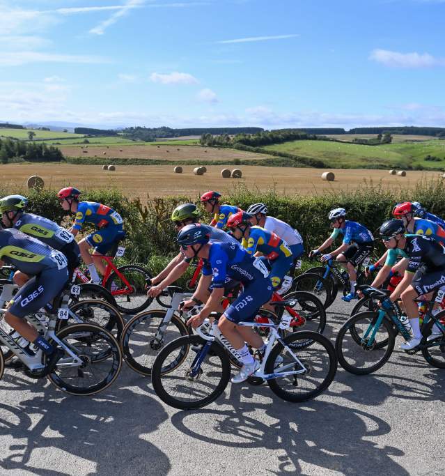 A group of cyclists rides past the camera during the Tour of Britain race, with rolling hills in the background.