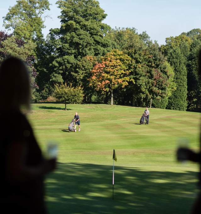 A golfing green with trees in the background and people holding drinks in the foreground
