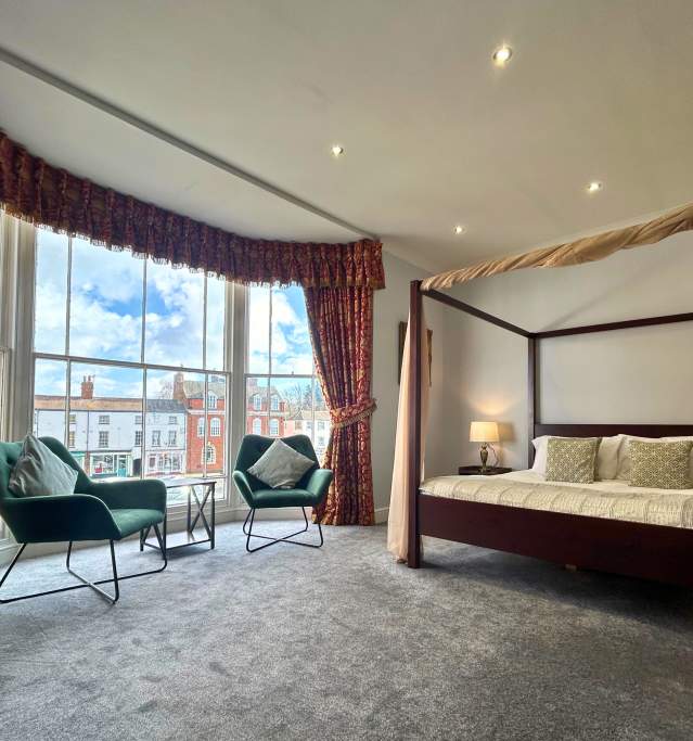 a bed and two chairs in a hotel room by a bay window overlooking a town square