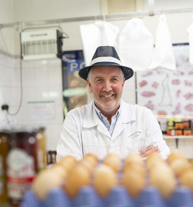 A man in butchers clothing looking at camera surrounded by butchers products