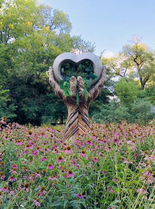 A large sculpture with hands that hold a heart. Pink flowers surround the sculpture.