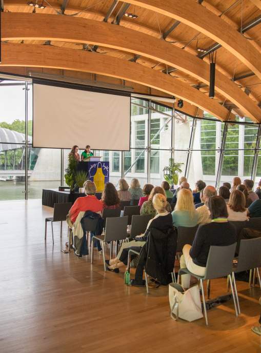People sitting in a Crystal Bridges Museum of American Art great hall. The room has glass windows and wooden beams on the ceiling.