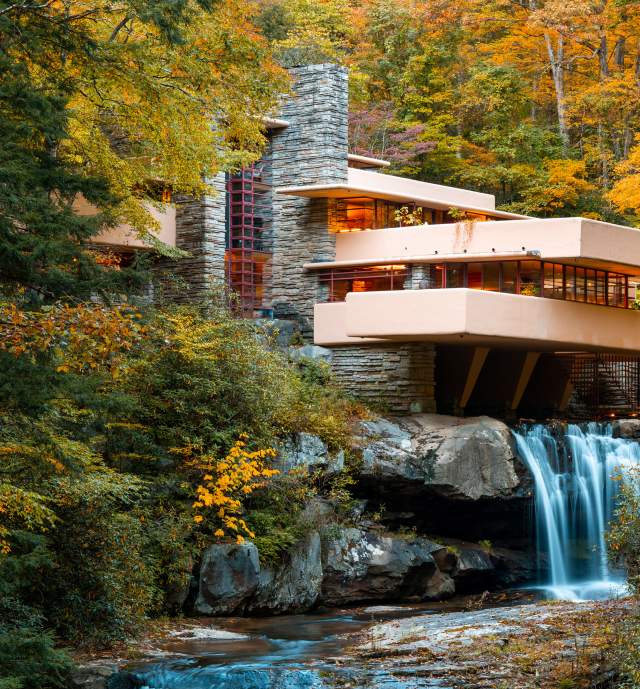 Iconic architectural building Fallingwater sits atop a waterfall with fall foliage surrounding.