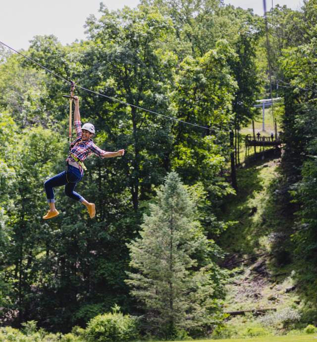 Woman ziplines down mountainside with pine trees in the background
