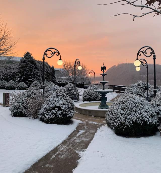 A quaint fountain in a park with snow covered ground and greenery surrounding and a sunset in the background at Danville's Riverfront