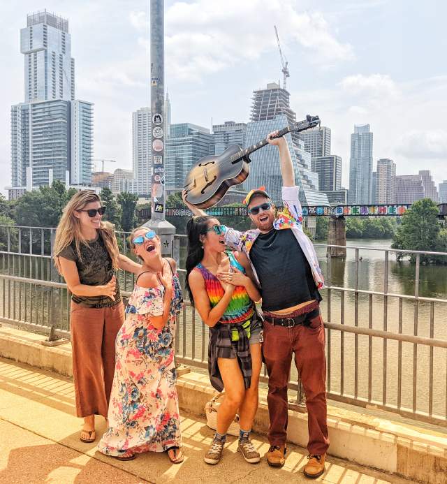 Four people posing on a bridge in front of Austin downtown skyline. Left to Right: woman grins at women beside her, woman places hand over heart and looks towards camera, woman places hands over her heart, man raises a guitar over his head