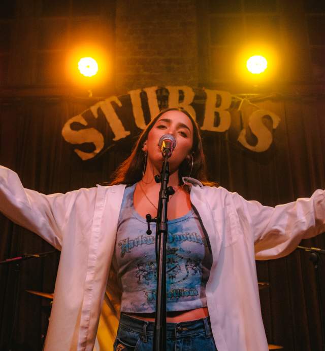 Woman standing in front of a microphone, singing with arms out, a large sign saying "STUBB'S" with pink and yellow neon lights is above her head.