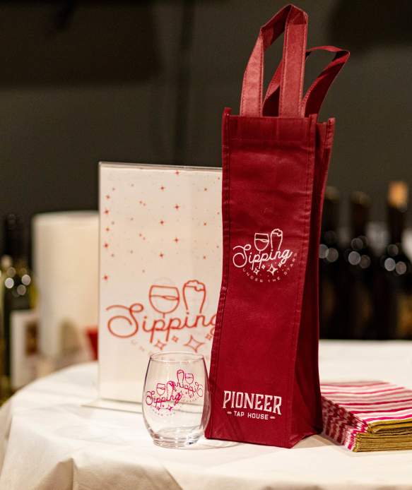 Photo of Sipping Under the Stars gear which includes a small wine glass and tote bag. Wine bottles can be seen in the background.