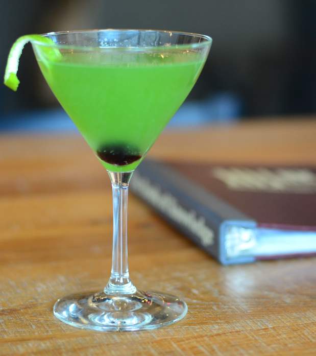 Bright green cocktail in a martini glass with cherry at borrom and lime twist garnish.