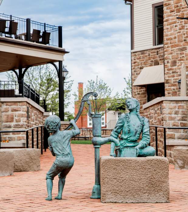 Bronze sculpture of a child pumping water for a seated Native American woman in a brick plaza surrounded by historic buildings.