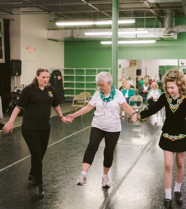 Older adults and a young Irish dancer hold hands in a circle during a lively dance demonstration in front of an audience.