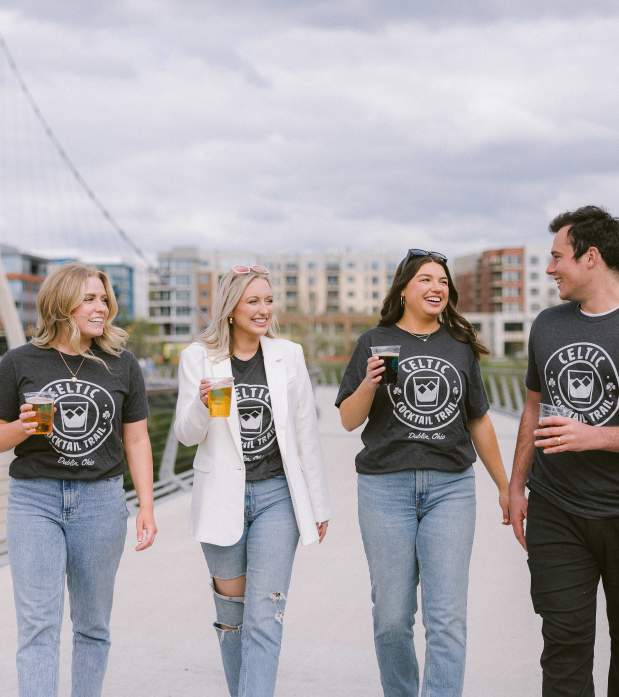A group of friends walking across the Dublin Link pedestrian bridge with DORA cups in Celtic Cocktail Trail t-shirts