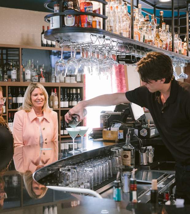 A bartender in a black shirt pours a green cocktail into a martini glass for two smiling women seated at a sleek bar with hanging glassware and liquor bottles in the background.