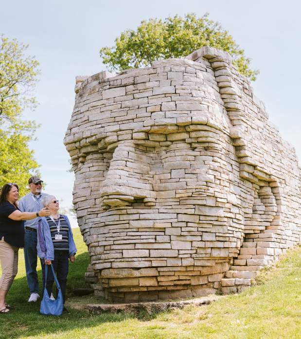 Three older adults admire a large stone sculpture of a face at the top of a grassy hill on a sunny day.
