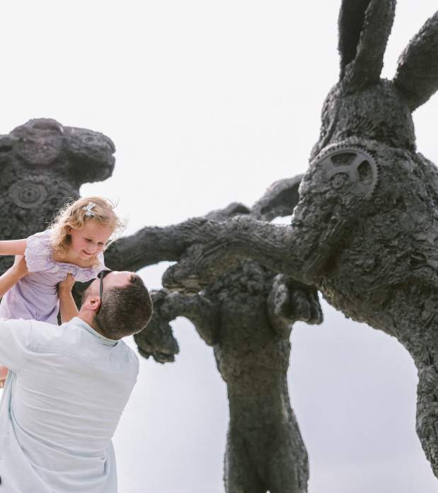 A father lifting his smiling daughter in front of large, textured rabbit sculptures in a whimsical outdoor setting, under a bright sky.