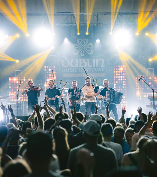 A lively concert at the Dublin Irish Festival featuring the band Skerryvore performing on a brightly lit stage with violins, bagpipes, and an accordion. The crowd is cheering with hands raised in excitement.