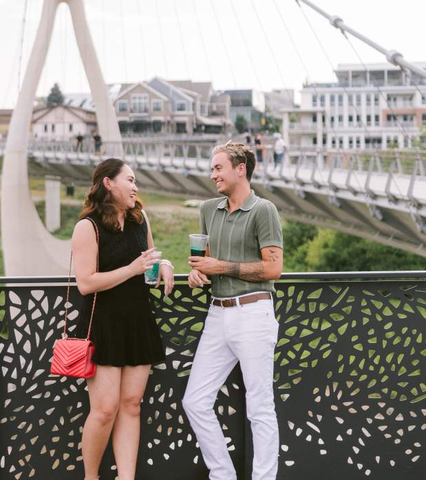 A young couple enjoying drinks and laughing on a decorative bridge railing with the Dublin Link pedestrian bridge and downtown Dublin, Ohio, in the background.