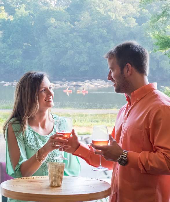 A man and a woman sharing wine together with a lake and a person walking their dog in the background