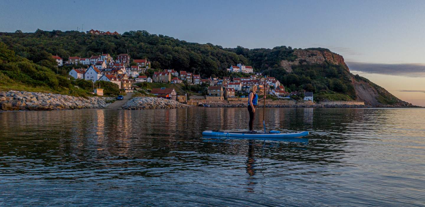 A paddle boarder heads out from runswick bay, the town reflected as a big smear across the shimmering sea