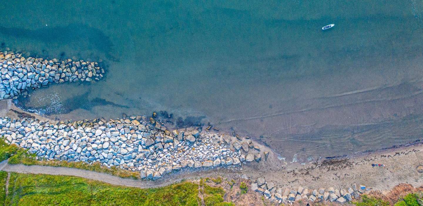An aerial shot of the shoreline, the huge granite boulder wall looking like mere gravel from this height