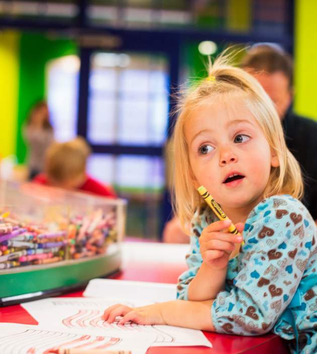 Little girl at a crafts table holding a crayon in her hand about to draw on a piece of paper.