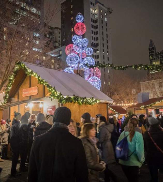People bundled up at the People's Gas Holiday Market in Pittsburgh at night with holiday lights on the vendor cottages.