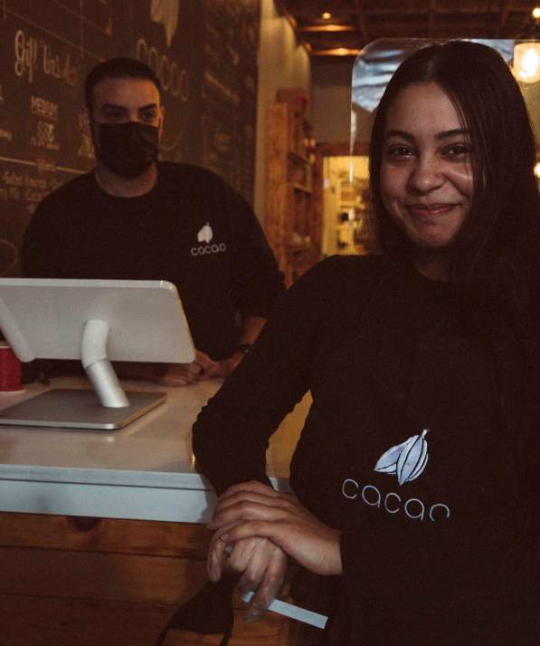 Woman leaning on counter in front of man wearing a cloth mask working behind a register at Cacao