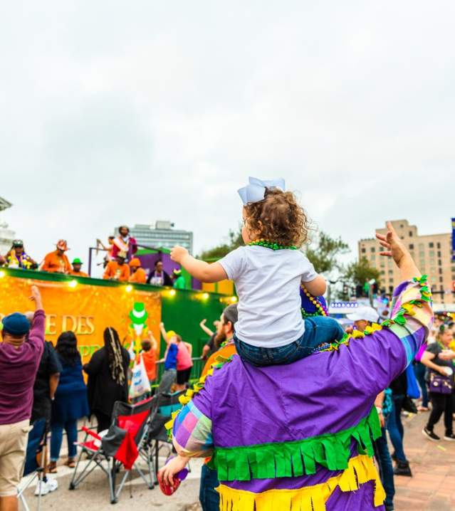 little girl sits on father's shoulders and catches beads in a crowd at a Mardi Gras parade