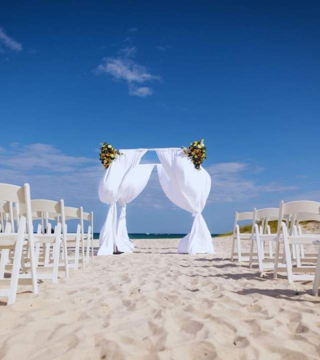 A wedding arch set up on the beach with white folding chairs lined up on either side of an aisle leading tot he arch