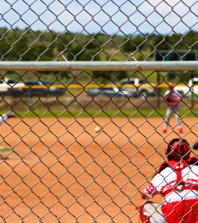 View from behind the fence  of a pitch for a baseball game with a team dressed in red and one in white at bat.