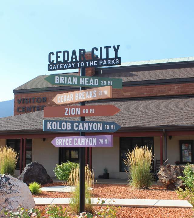 A brightly colored wayfinding sign pointing to nearby national parks and monuments standing in front of the Cedar City Visitor Center.