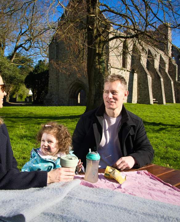Family enjoying a picnic on a sunny day at a picnic table in front of Grey Abbey, County Down.