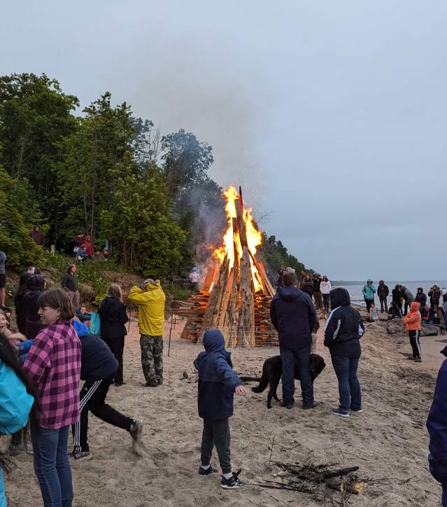 a crowd gathers around an enormous beachside bonfire