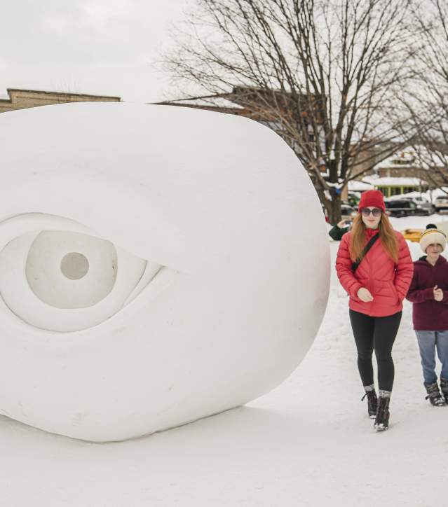 two parents and two children walk together in snow gear next to a snow statue of a human eye taller than an average person