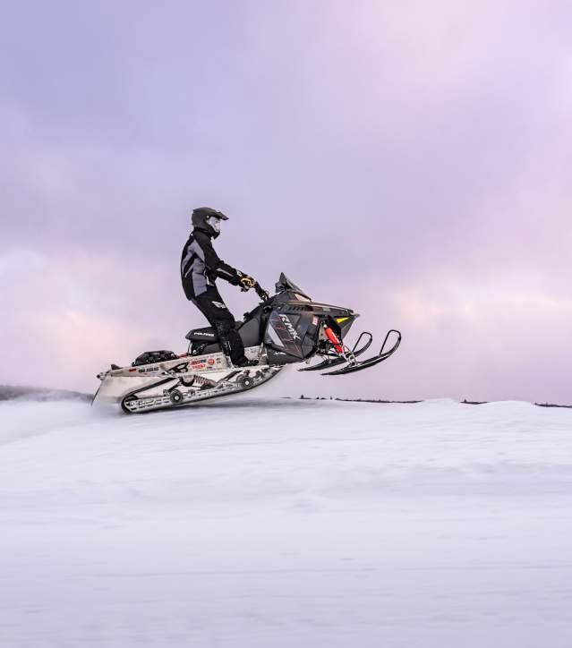 A rider makes a small jump with his snowmobile with a purple sky behind.