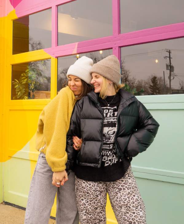 Two women wearing beanies and coats hold hands and stand in front of a colorful mural.