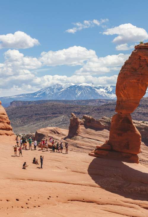 Delicate Arch pictured with a group of about 15 people looking at it. In the background, the La Sal Mountains are topped with snow.