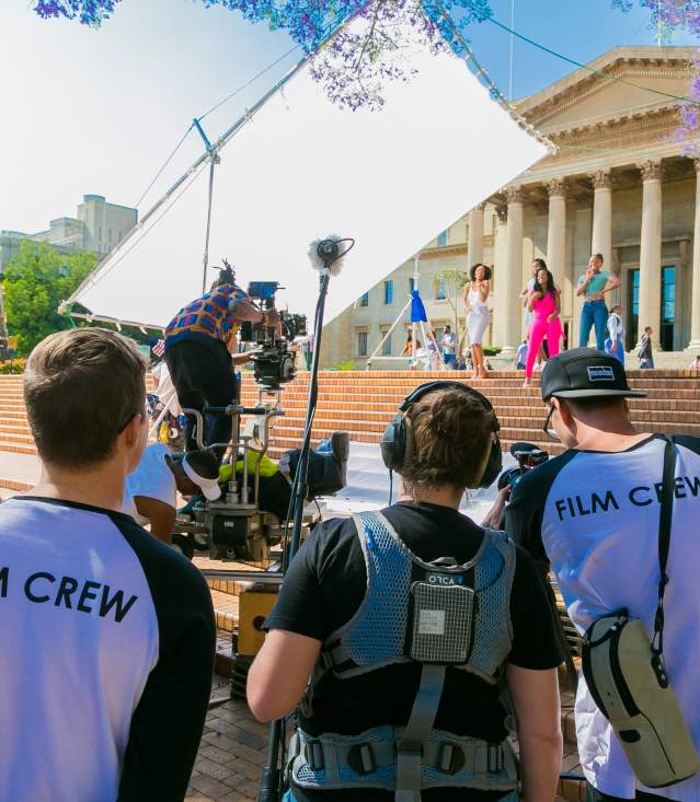 Production crew filming girls singing on the steps in front of a building with columns.