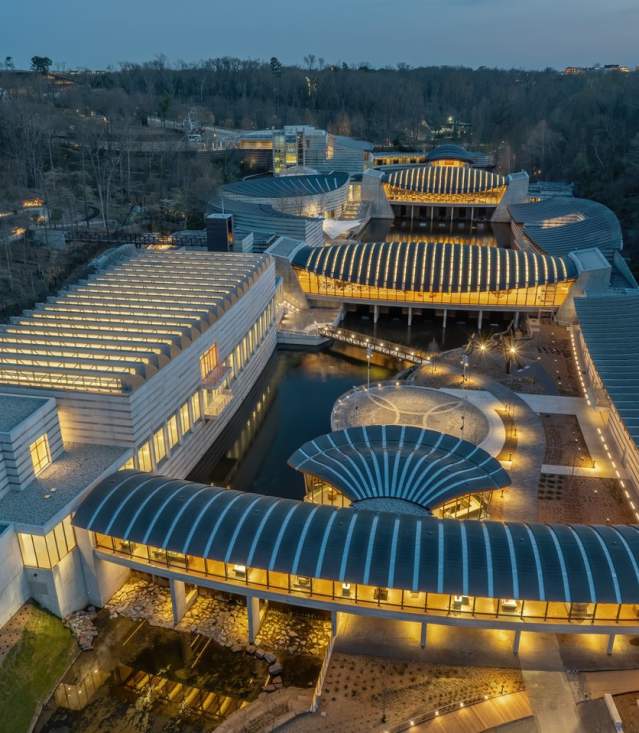 Aerial view of a modern architectural complex with flowing lines, illuminated pathways, and surrounding greenery at dusk. The design features multiple interconnected buildings and water elements.