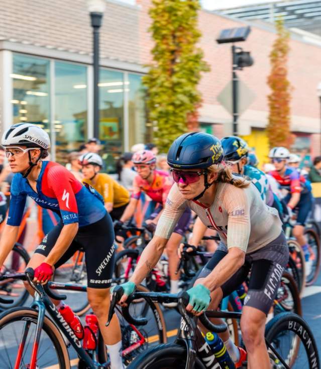 A group of cyclists in colorful jerseys races through a city street lined with trees and buildings, showcasing an energetic urban event.