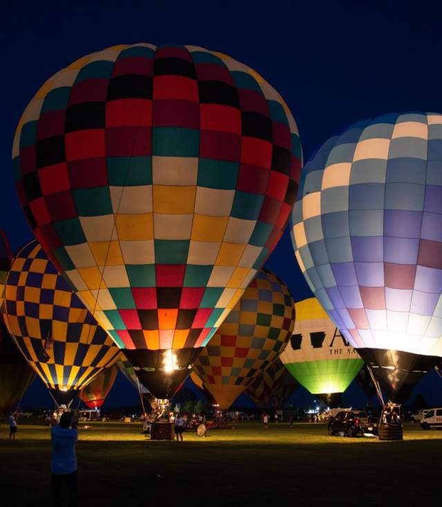 Colorful hot air balloons illuminated at night, set against a dark sky. Spectators gather around, creating a vibrant atmosphere.