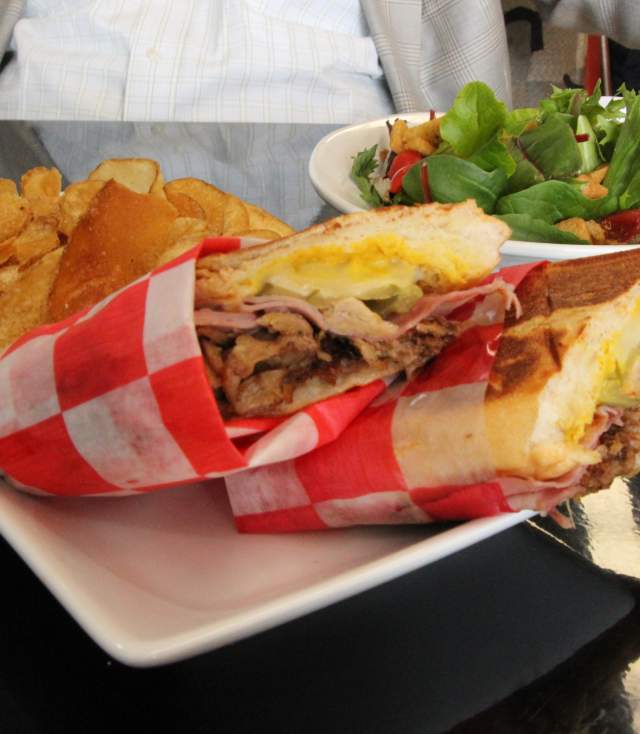 Plate with a toasted sandwich wrapped in red-and-white checkered paper, served with golden potato chips and a side salad of fresh greens.