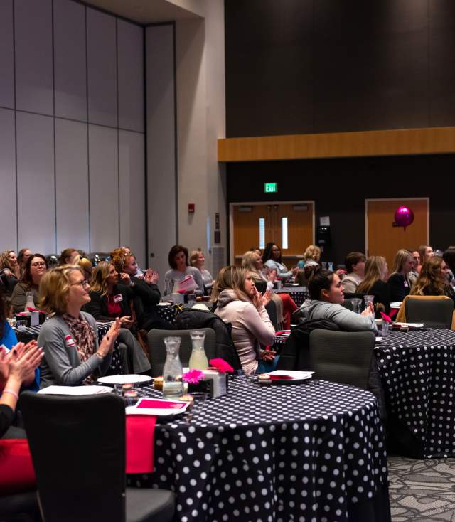 Dozens of women sit at round tables in a large room applauding the stage at the annual Lead Her Forward event
