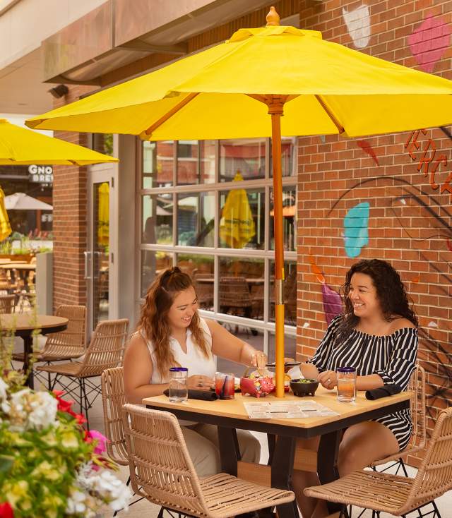 two women eating at an outdoor table at a restaurant sitting under a yellow umbrella with a mural on the wall behind them