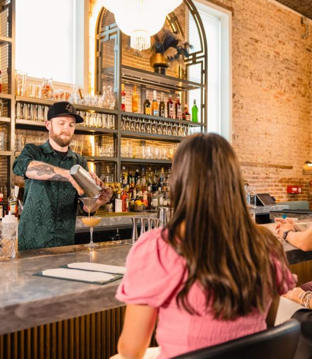 A bartender with tattoos pours a cocktail into a glass at Lady Slipper bar, with a backdrop of a well-stocked bar shelf and exposed brick walls, as guests seated at the bar watch with interest.