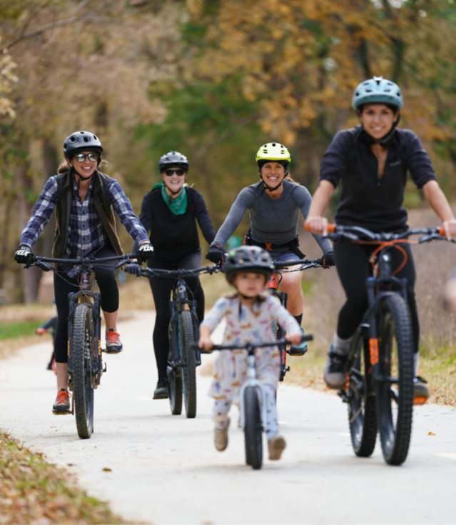 A mix of women and children riding mountain bikes cruise on a paved trail in colorful bike helmets.