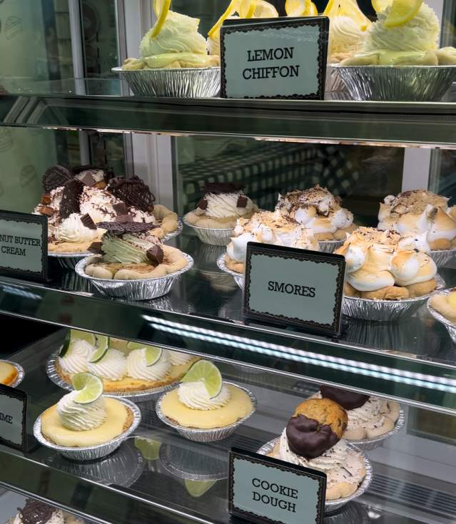 Display case filled with an assortment of Gooseberry Handmade Pies, featuring Lemon Chiffon, S'mores, Key Lime, and Cookie Dough pies, each topped with themed garnishes and placed under descriptive signs.