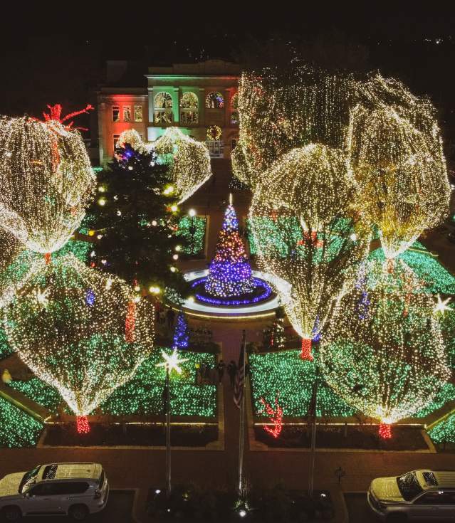 Drone photo of holiday lights on the square. Lights on the ground are green, tree stumps are wrapped in red, and branches and leaves are wrapped in white lights.