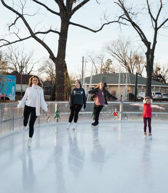 Four people skate on an outdoor ice rink surrounded by trees and buildings. Colorful decorations hang around the rink, enhancing the festive atmosphere.