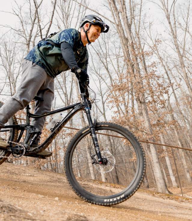 A mountain biker navigates a dirt trail, surrounded by bare trees in a wooded area. The rider wears a helmet and sports gear, showcasing an active outdoor experience.
